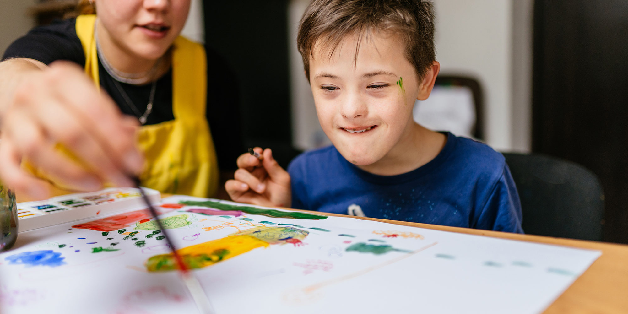 Young female teacher explain, talk at desk with a down syndrome schoolboy. Color painting for disabled kids, child who are down syndrome and student teacher, development of children with spesial needs